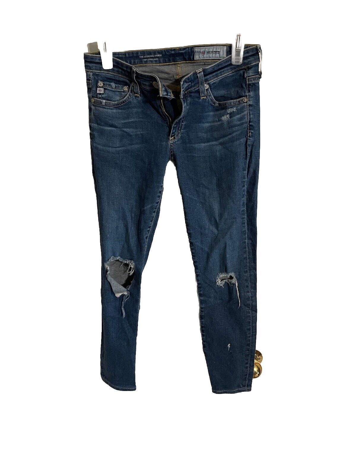 Distressed dark wash AG legging ankle jeans with front and knee rips hanging on white clips