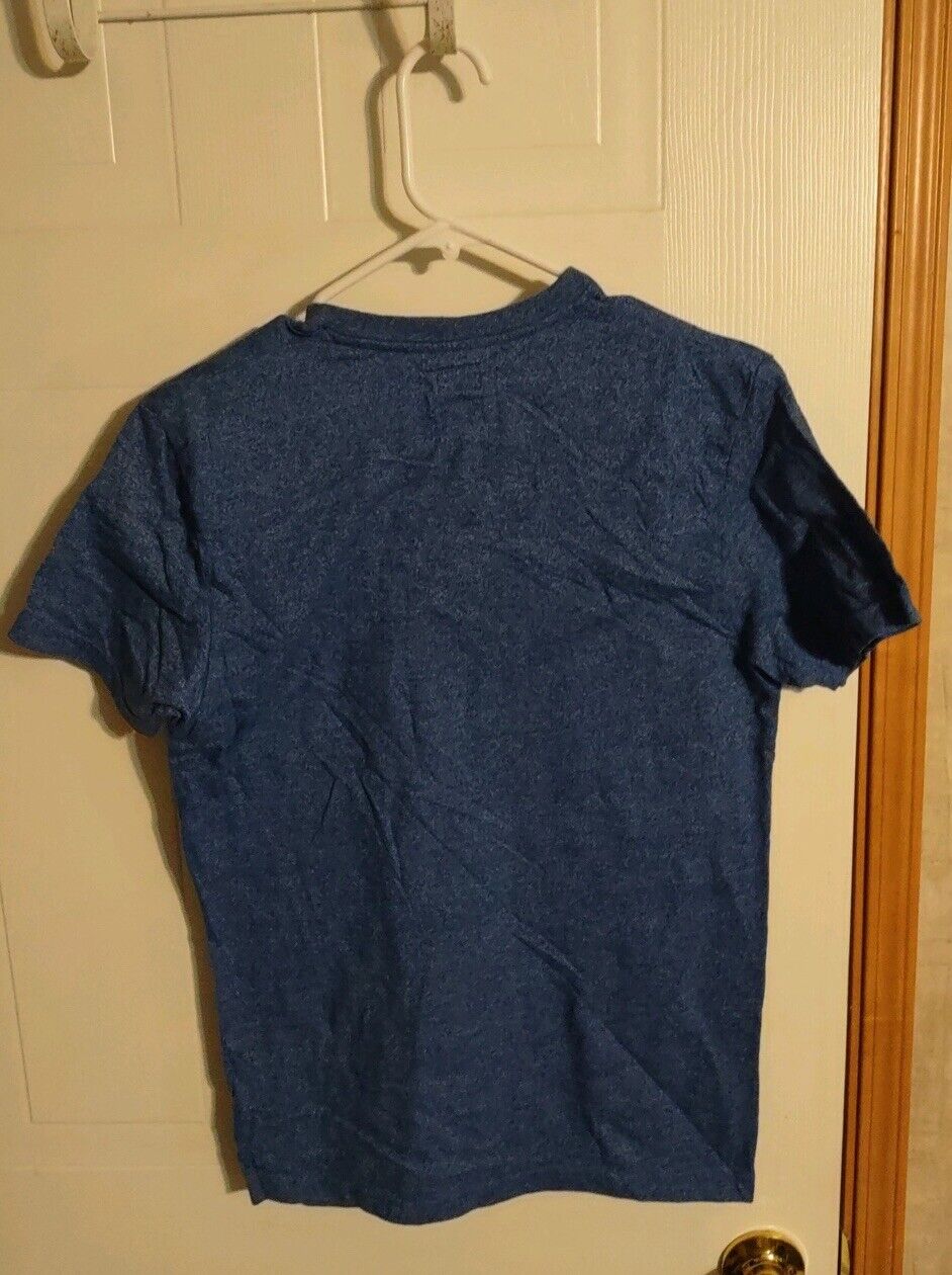 Alphabeta blue t-shirt small hanging on a white plastic hanger against a beige door background