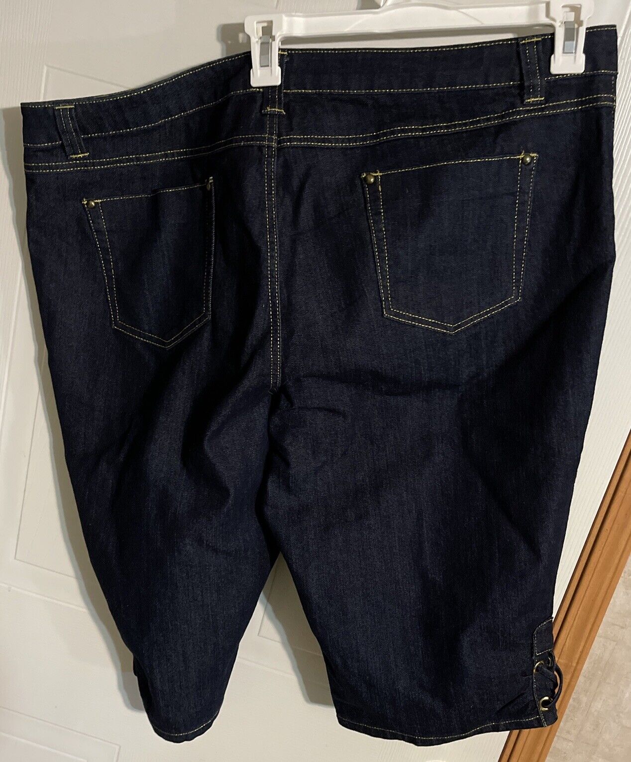 Back view of dark denim Ashley Stewart jorts 22W with yellow stitching and pockets hanging on a white hanger