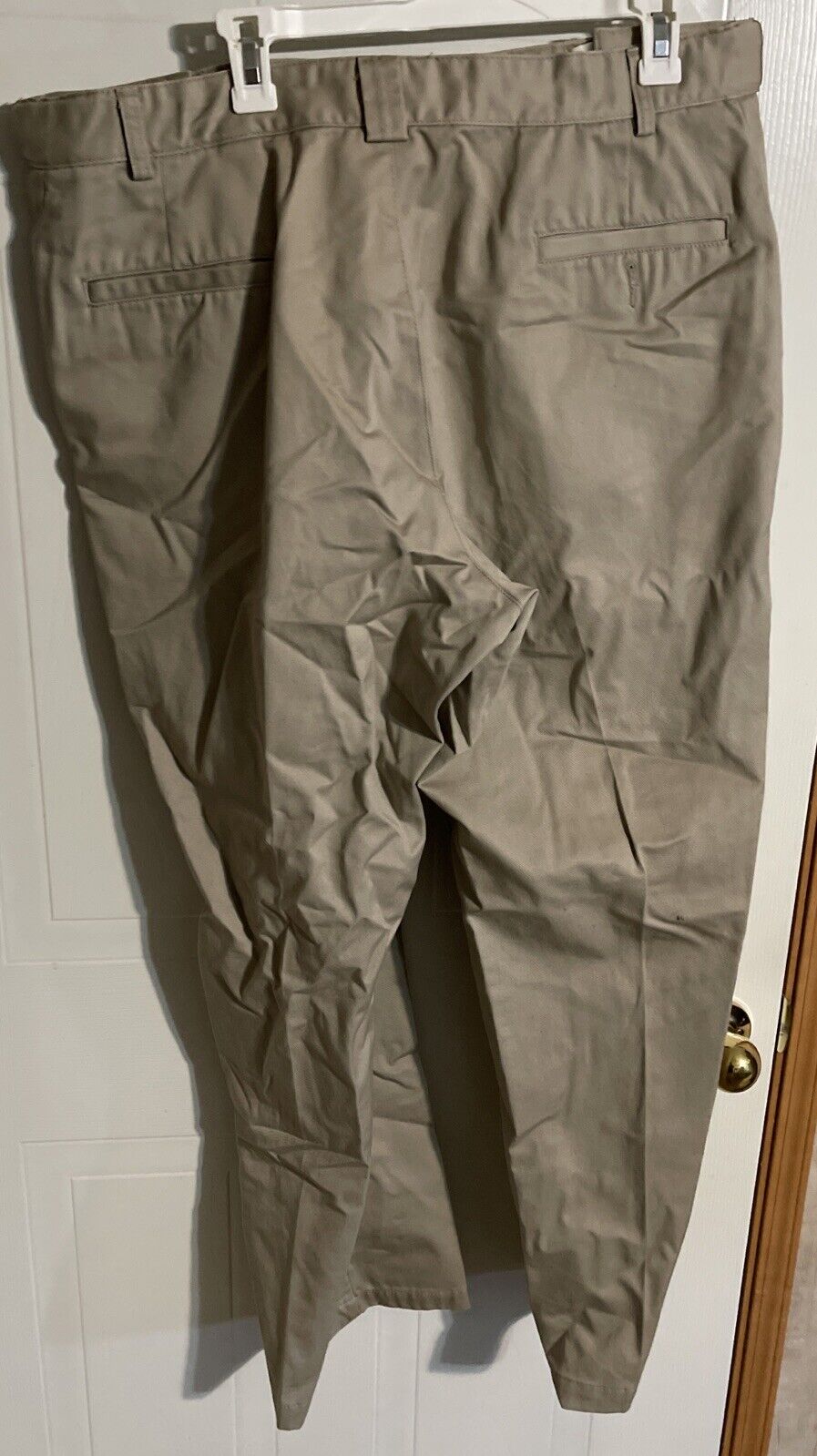 Beige men's tactical work pants hanging on a white door with belt loops and back pockets