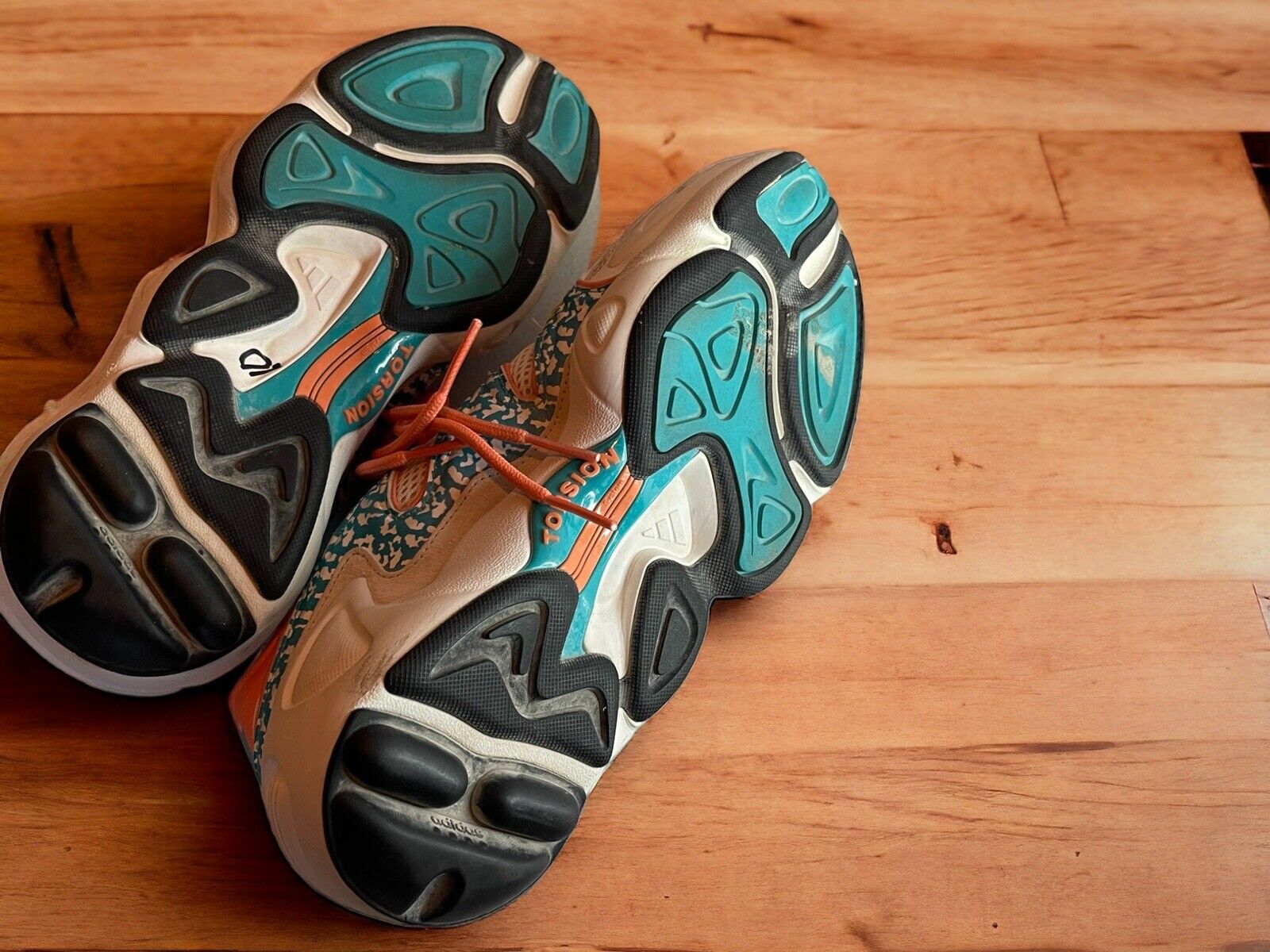 Bottom view of Adidas Originals FYW S-97 sneakers showing black and teal sole design on wooden floor