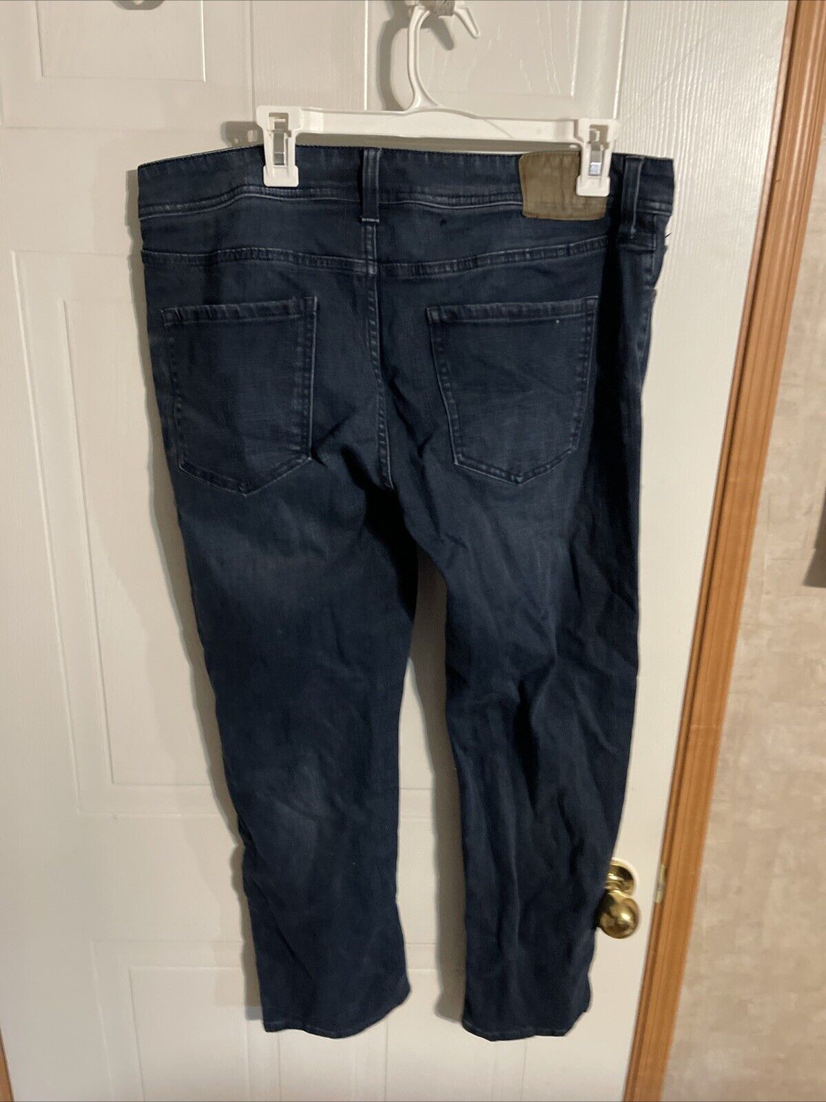 Back view of dark blue American Rag CIE jeans hanging on white hanger against white door