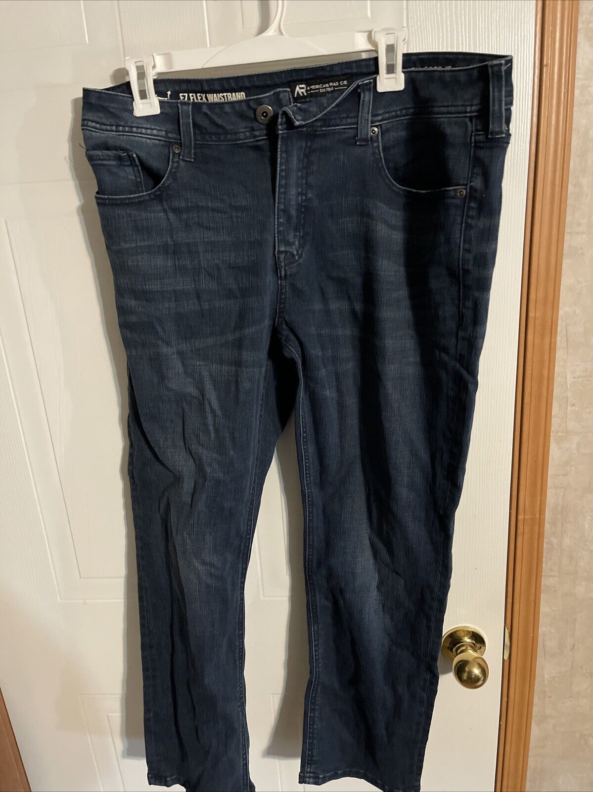 Dark wash American Rag CIE jeans hanging on a white hanger against a white door