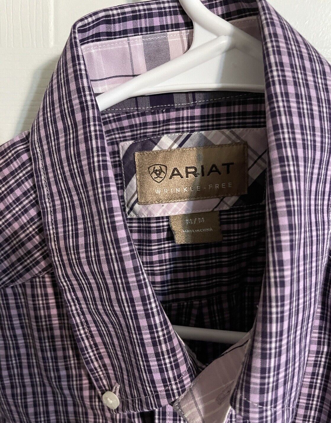 Purple and black plaid Ariat wrinkle free shirt hanging on a white hanger with visible brand label