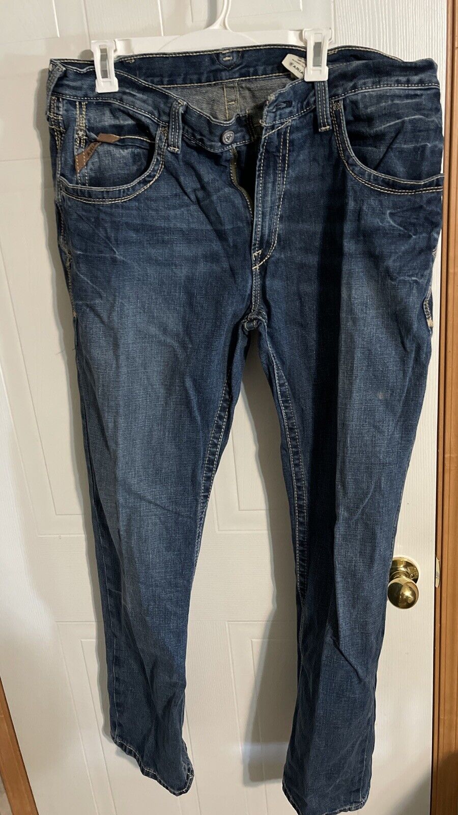 Ariat jeans 38x34 in dark blue denim hanging on white hanger against a door background