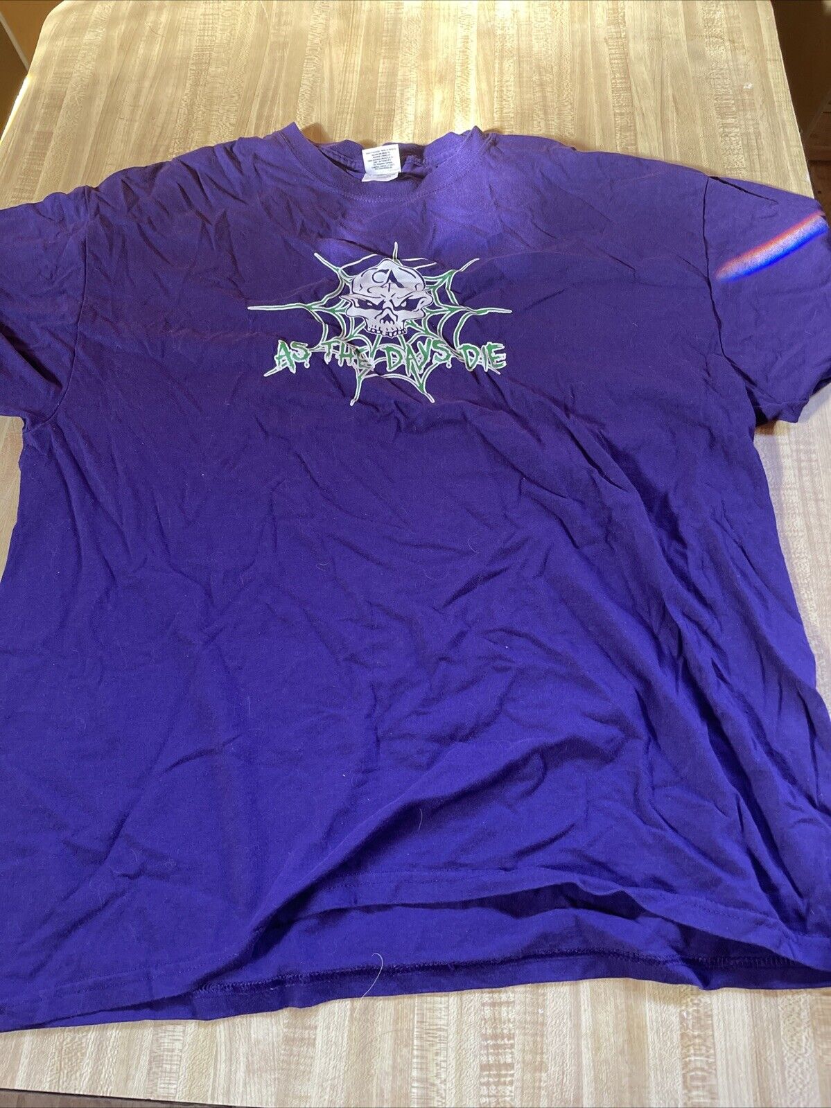 Purple As The Days Die band shirt with skull and web graphic on front laid flat on wooden surface