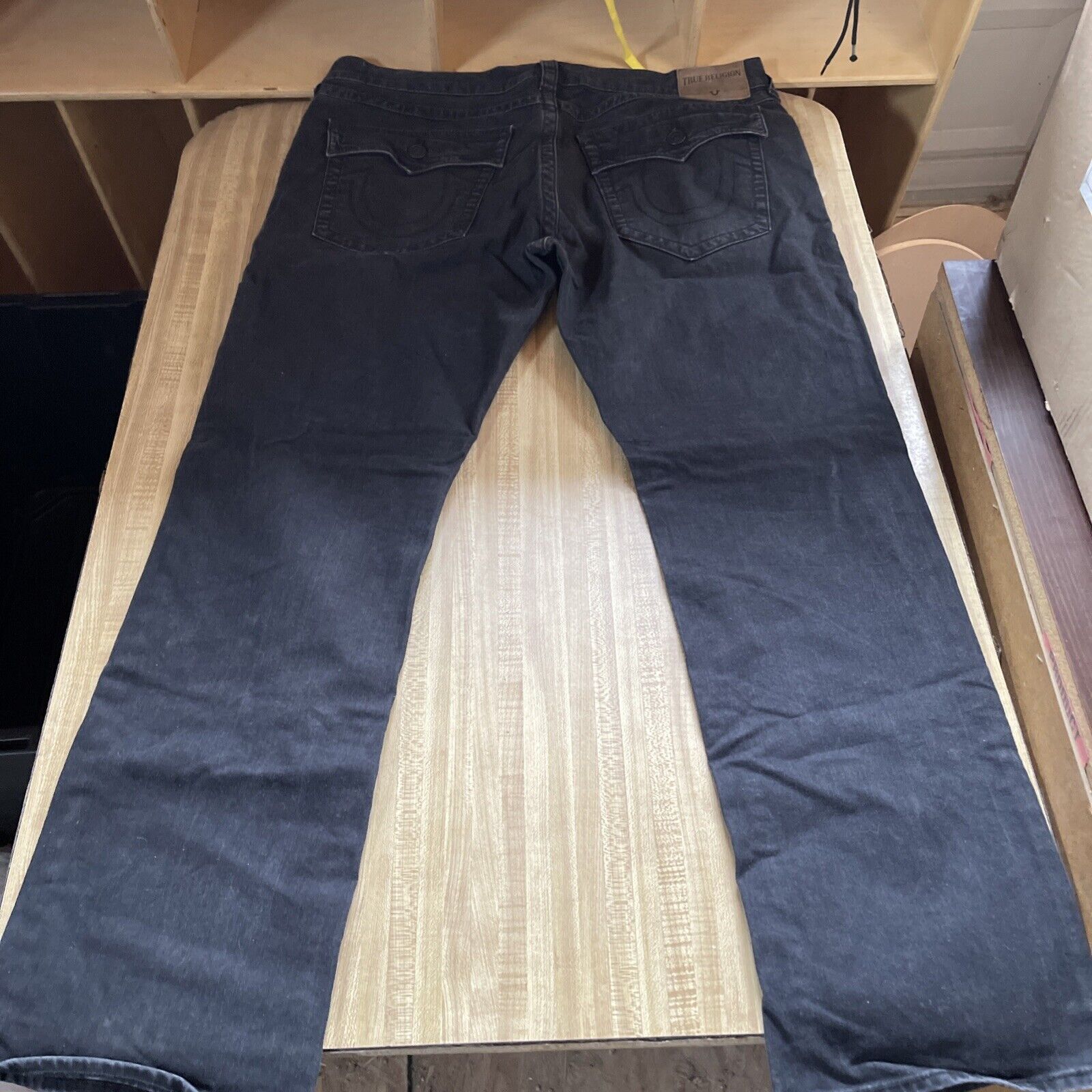 Black denim True Religion Ricky jeans 40x34 laid flat on wooden surface showing back pockets and label