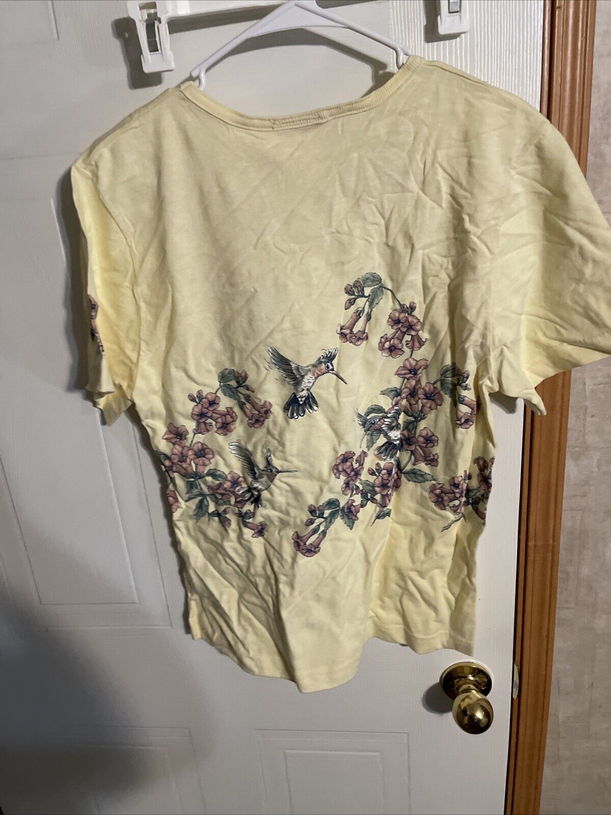 Cream yellow vintage art unlimited t shirt with hummingbird and floral design hanging on white door